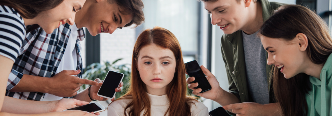Abbildung ein rothaariges Mädchen wird von vier Jugendlichen umkreist, sie schaut traurig, während die anderen vier lachen und auf deren Handys zeigen.